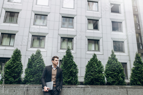 Portrait of a handsome businessman in a stylish suit who stands near a business center outdoor with a laptop © Kateryna