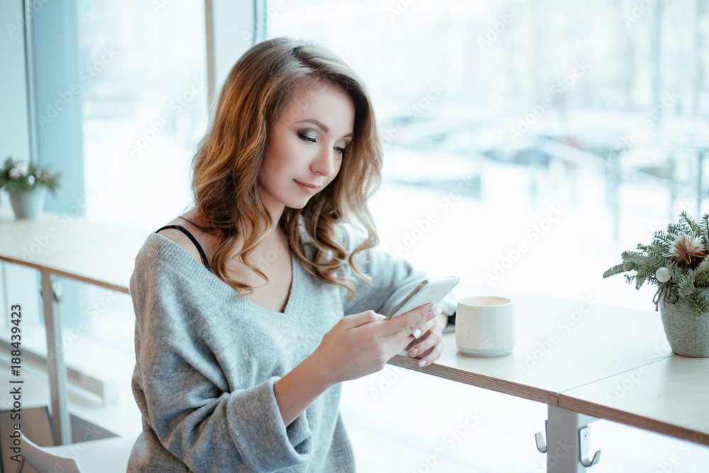Beautiful happy pretty young woman sitting in a cafe with mobile phone. White and blue colors. cup of coffee and decorative flowers on a table. Beautiful happy pretty young woman sitting in a cafe with mobile phone. White and blue colors. cup of coffee and decorative flowers on a table.