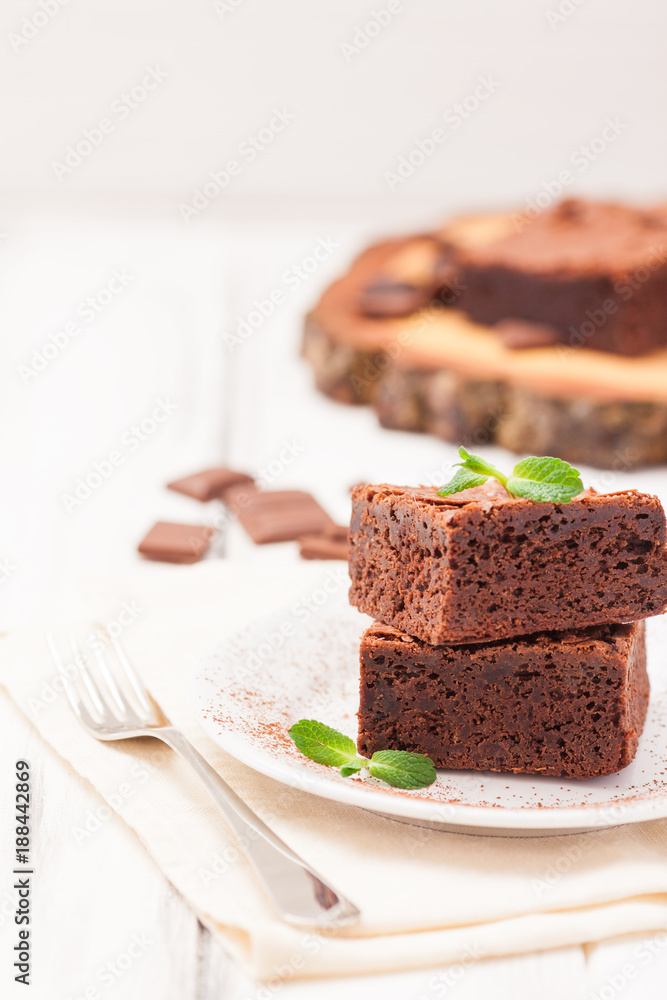Chocolate brownie square pieces in stack on white plate decorated with mint leaves and cocoa powder on white vintage wooden background. American traditional delicious dessert. Close up photography