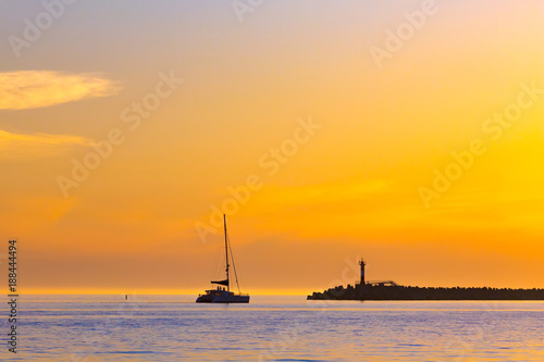 Summer bright orange sunset with beautiful clouds and the silhouette of a sailing boat with a descended sail next to the lighthouse on the beach. Coastal seascape on the Black Sea, Sochi, Russia.