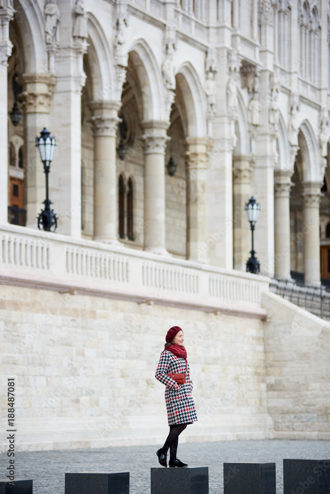 Woman stands against the backdrop of a powerful European architectural structure. She has a red beret and a handbag, a coat in tone. The concept of the autumn Woman stands against the backdrop of a powerful European architectural structure. She has a red beret and a handbag, a coat in tone. The concept of the autumn
