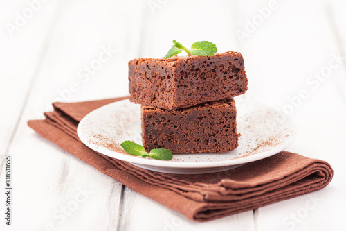 Chocolate brownie square pieces in stack on white plate decorated with mint leaves and cocoa powder on white vintage wooden background. American traditional delicious dessert. Close up photography