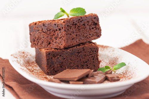 Chocolate brownie square pieces in stack on white plate decorated with mint leaves and cocoa powder on white vintage wooden background. American traditional delicious dessert. Close up photography