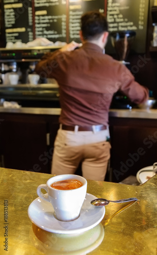 Typical Parisian cafe bar. Coffee cup on the shiny counter. Barista making a coffee  back view  at backgrounds. Selective focus on the foam in the cup. 