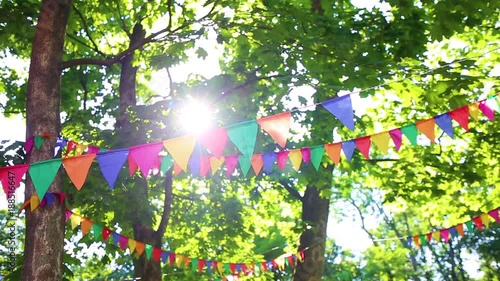 Colorful flags hanging on branches of trees as festive decoration for outdoor party. Bright summer sunshine among green leaves in background. Real time full hd video footage.