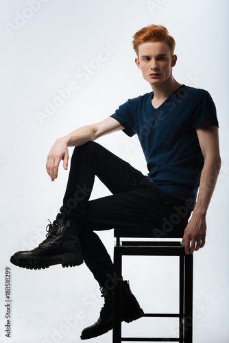 Seriousness. Good-looking humourless red-haired young man sitting on the chair and having his hand on his knee and wearing a black t-shirt and trousers and boots photo