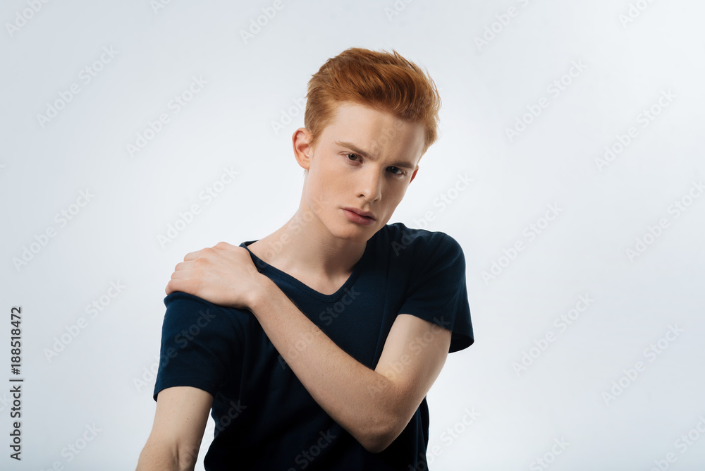 Meditative. Handsome meditative red-haired young man sitting and staring and having his hand on his shoulder