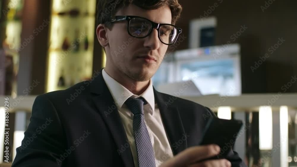Young handsome businessman in eyeglasses surfing the Internet on smartphone while sitting in restaurant Young handsome businessman in eyeglasses surfing the Internet on smartphone while sitting in restaurant