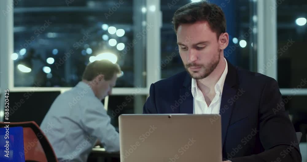 An employe in black suit in the evening office. Sitting at the laptop and checking documents. Late work overtime concept. An employe in black suit in the evening office. Sitting at the laptop and checking documents. Late work overtime concept.