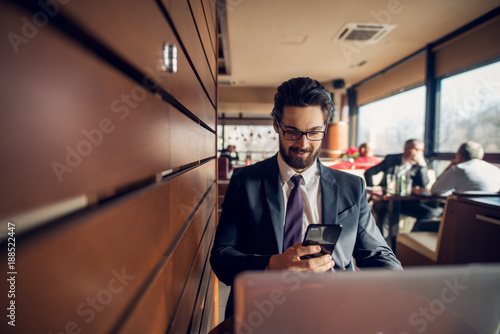 Satisfied stylish smiling handsome businessman in the suit reading a message from a mobile while sitting with a laptop next to the wooden wall in a cafe or restaurant. © dusanpetkovic1