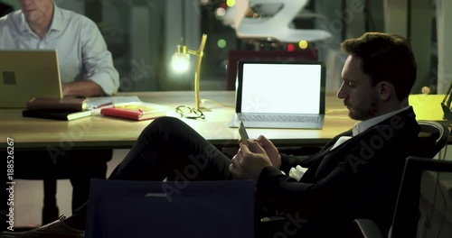 Business partners working late in office. One businessman is working on laptop, another one is typing message on phone. Late work overtime concept.