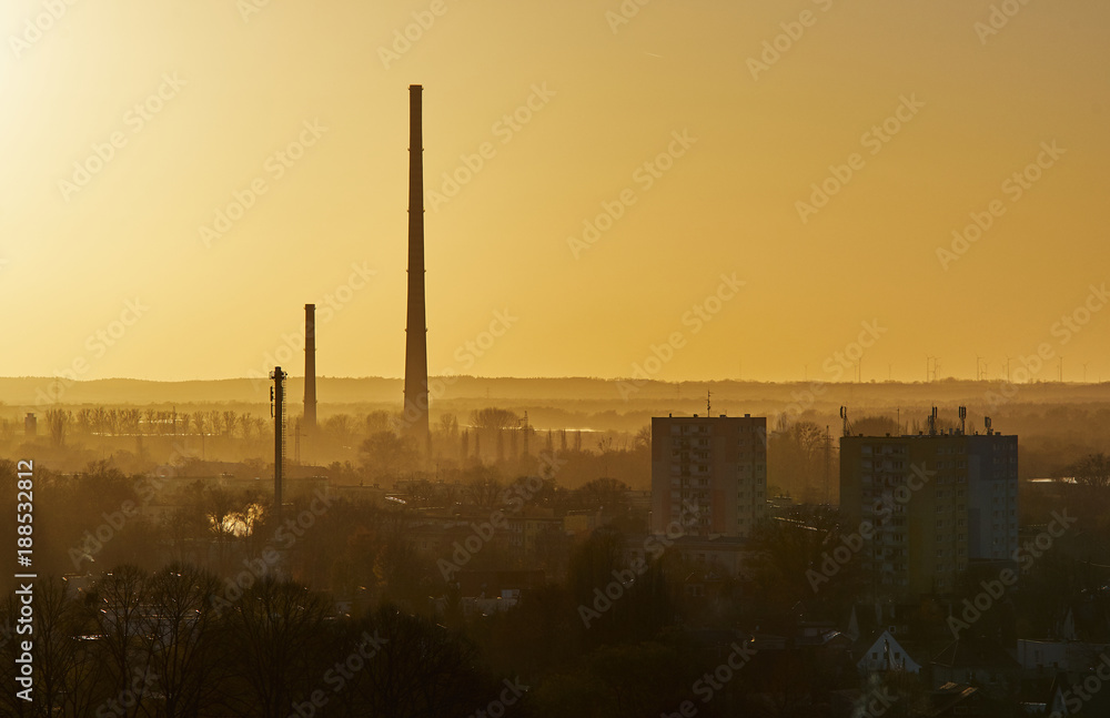 Skyscrapers and chimneys of the factory among trees, buildings and fog.