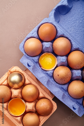Vertical closeup shot with two colorful cardboard containers of pink and violet color with chicken eggs,  two yolks visible, one egg painted golden. Easter concept