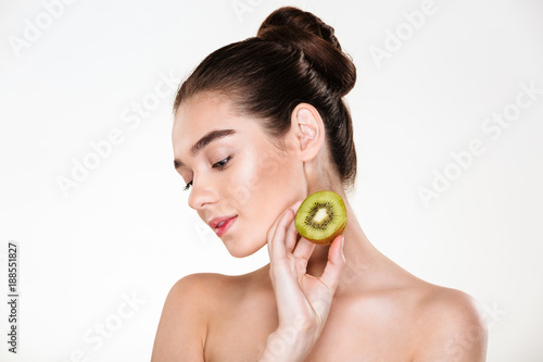 Picture of tender woman having clean skin posing isolated over white background with closed eyes holding ripe juicy kiwi in hand