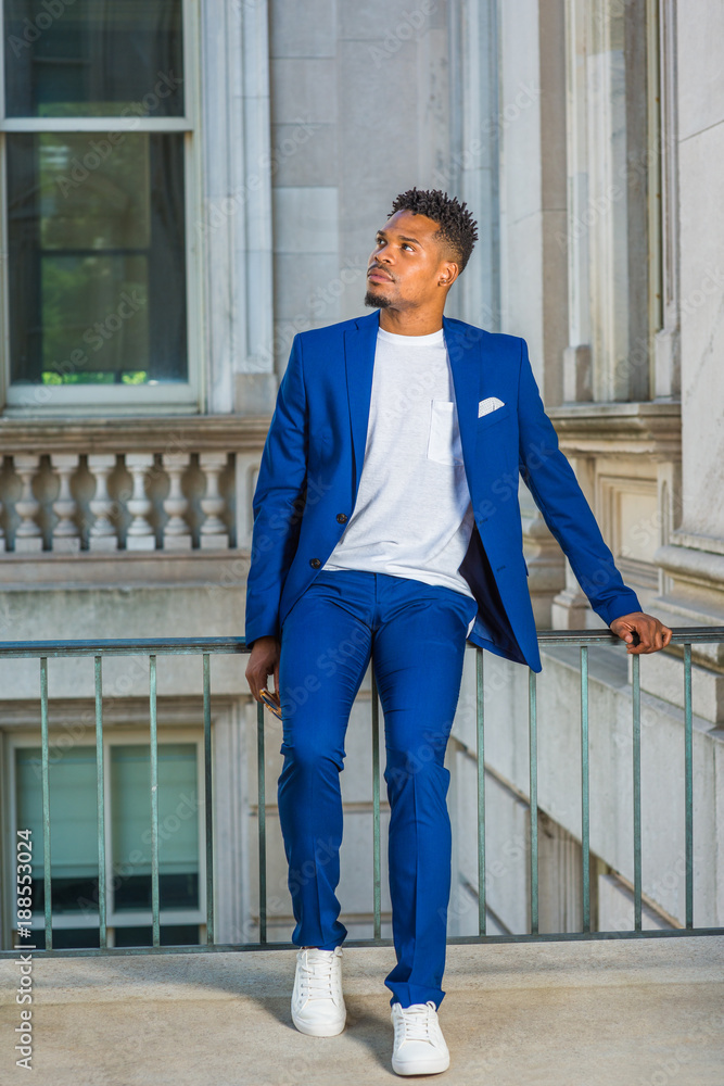 African American college student studying in New York, wearing blue suit,  white collarless shirt, sneakers, sitting on railing in vintage office  building on campus, taking break, looking up, thinking Stock Photo