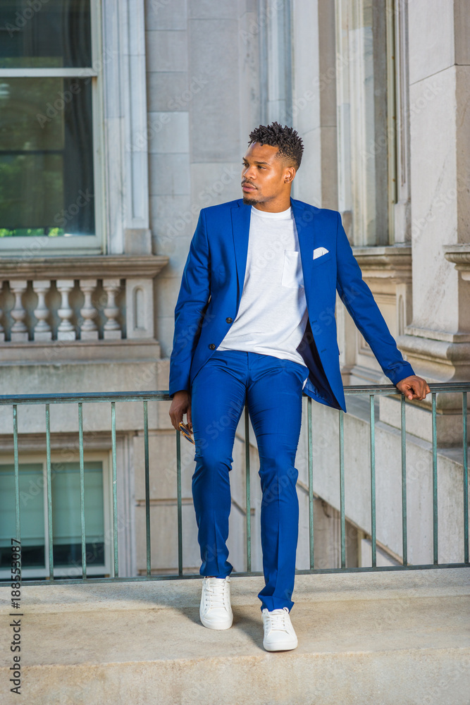 African American college student studying in New York, wearing blue suit,  white collarless shirt, sneakers, sitting on railing in vintage office  building on campus, taking break, looking away. Stock Photo | Adobe