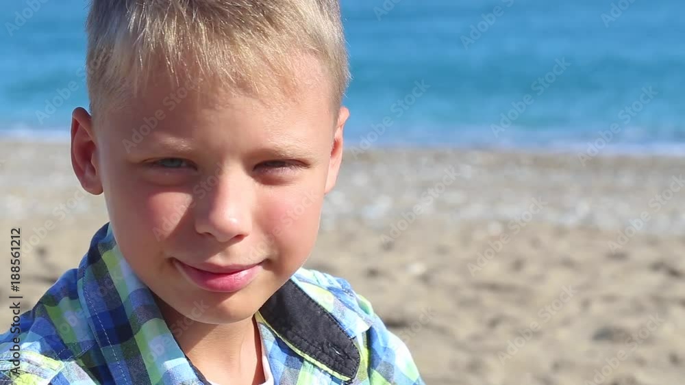 Closeup portrait of cute blond kid sitting isolated at blurry blue sea water background. White tan child puts thumb up at sandy sunny beach happily. Closeup portrait of cute blond kid sitting isolated at blurry blue sea water background. White tan child puts thumb up at sandy sunny beach happily.