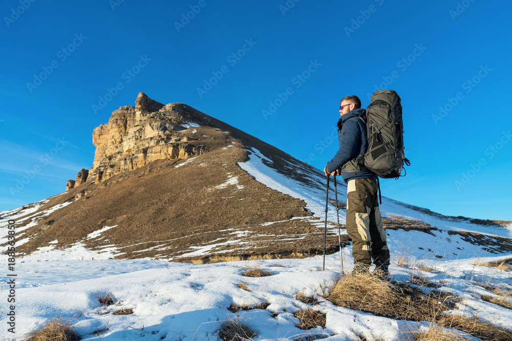 Backpacker with a large backpack and sticks ascends to the rock on sunset against the background of epic rocks in the winter season.