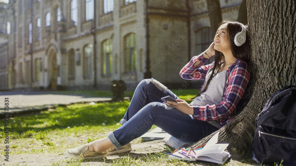 Female student sitting on grass, leaning against tree, listening to music, relax Female student sitting on grass, leaning against tree, listening to music, relax