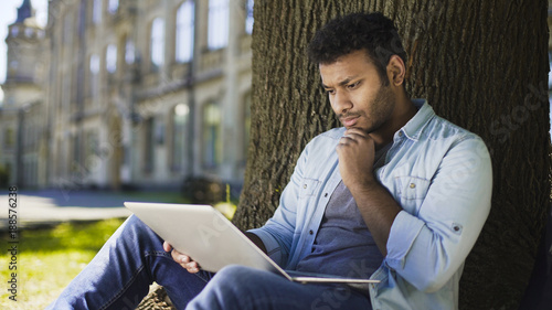 Mixed-race young man sitting under tree, looking at laptop, important letter