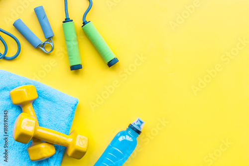 Fitness background. Equipment for gym and home. Jump rope, dumbbells, expander, mat, water on pastel yellow background top view copy space