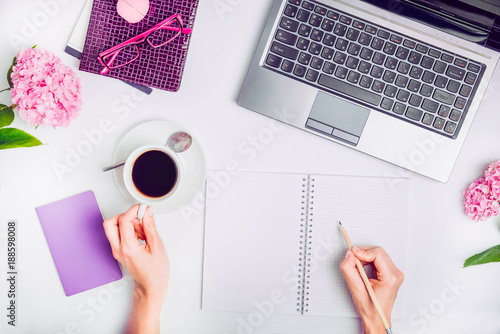 Workspace with laptop, girl's hands writing in notebook, glasses, cup of coffee and wisteria flowers on white background. Top view feminine office table desk. Freelancer working place. photo