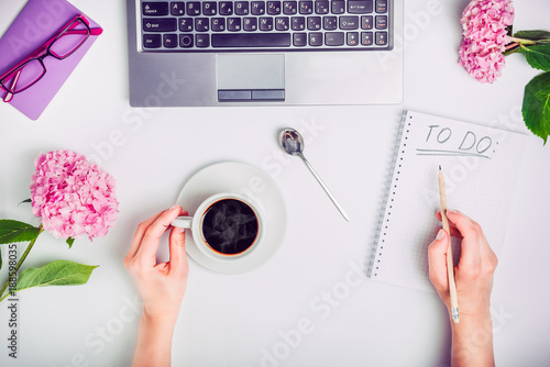 Day Planning - female hands with cup of coffee and pencil write To do list on the white working office desk with laptop, notebook, glasses, and wisteria flowers. Freelance. photo