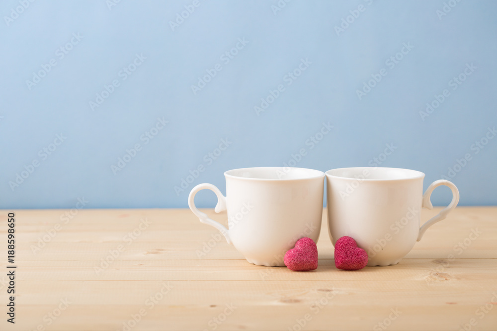 Valentines day concept, mugs cups of coffee or tea for two lovers honeymoon wedding morning surprise breakfast, pink heart sugar candies, blue background pastel colors copy space, light wooden table 