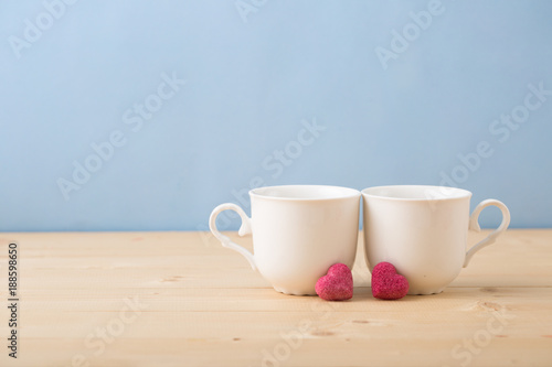 Valentines day concept, mugs cups of coffee or tea for two lovers honeymoon wedding morning surprise breakfast, pink heart sugar candies, blue background pastel colors copy space, light wooden table 