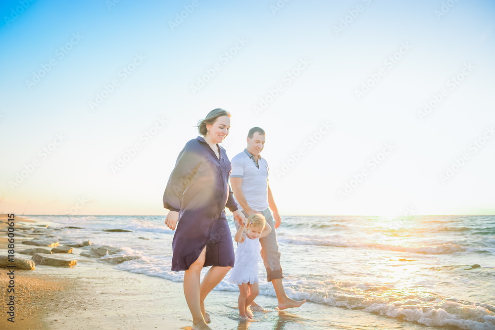 Happy family of three - pregnant wife, father and daughter having fun walking on beach at sunset. Family traveling concept. Backlight, soft selective focus. Copy space.