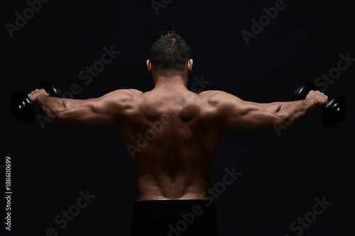 Muscular bodybuilder doing exercises with dumbbell over black background.Strong athletic man shows body,abdominal muscles,biceps and triceps.Work out,gaining weight,pumping up muscles with dumbbells.