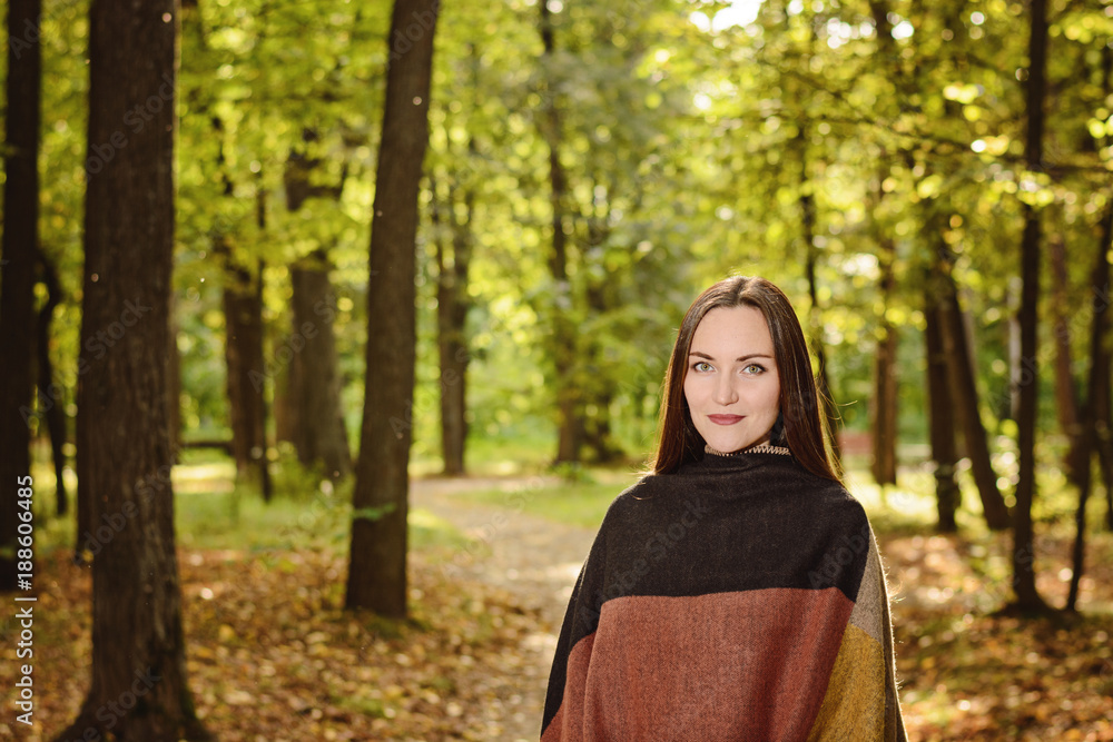 Portrait of a young woman brunette in a wool poncho Standing on a footpath in the green Park in the background in the rays of the bright sun Portrait of a young woman brunette in a wool poncho Standing on a footpath in the green Park in the background in the rays of the bright sun