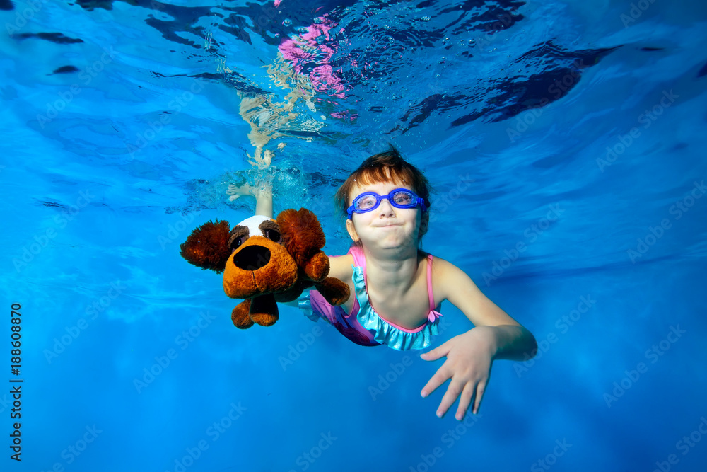 Φωτογραφία Happy little girl swims underwater in the pool, holding a toy dog in hand, looking at camera and smiling