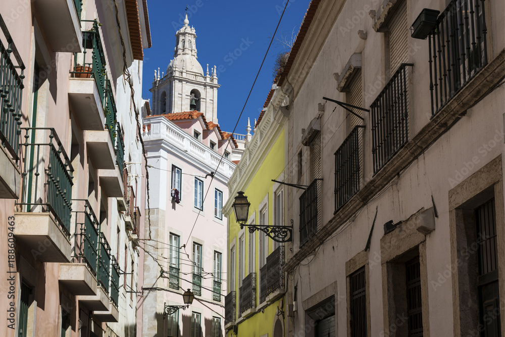 View of a narrow street and buildings with a tower of the Sao Vincente de Fora church on the backrgound, in the historic neighborhood of Alfama in Lisbon, Portugal; Concept for visit Lisbon