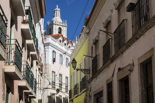 View of a narrow street and buildings with a tower of the Sao Vincente de Fora church on the backrgound, in the historic neighborhood of Alfama in Lisbon, Portugal; Concept for visit Lisbon
