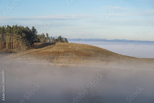 Fototapeta Naklejka Na Ścianę i Meble -  A small hill rising just above the thick fog on a sunny afternoon in the foothills of the Carpathian mountains