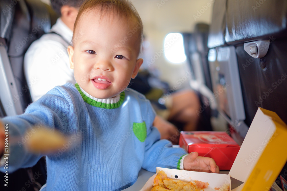 Cute little Asian 18 months / 1 year old toddler baby boy child wearing blue sweater eating food during flight on airplane. Flying with children, Happy air travel with kids & little traveler concept Cute little Asian 18 months / 1 year old toddler baby boy child wearing blue sweater eating food during flight on airplane. Flying with children, Happy air travel with kids & little traveler concept