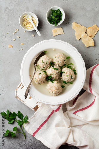 Homemade chicken matzo ball soup with parsley and garlic in simple white ceramic plate on a gray stone or concrete background. Traditional Jewish passover dish. Selective focus. Top view.