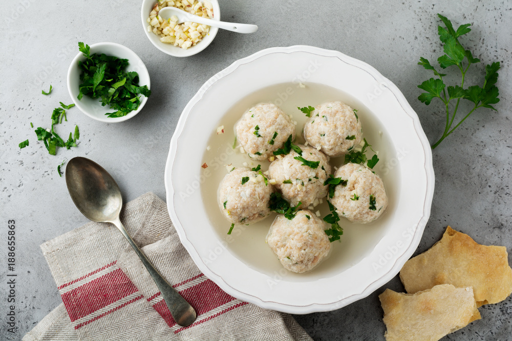 Homemade chicken matzo ball soup with parsley and garlic in simple white ceramic plate on a gray stone or concrete background.  Traditional Jewish passover dish. Selective focus. Top view.