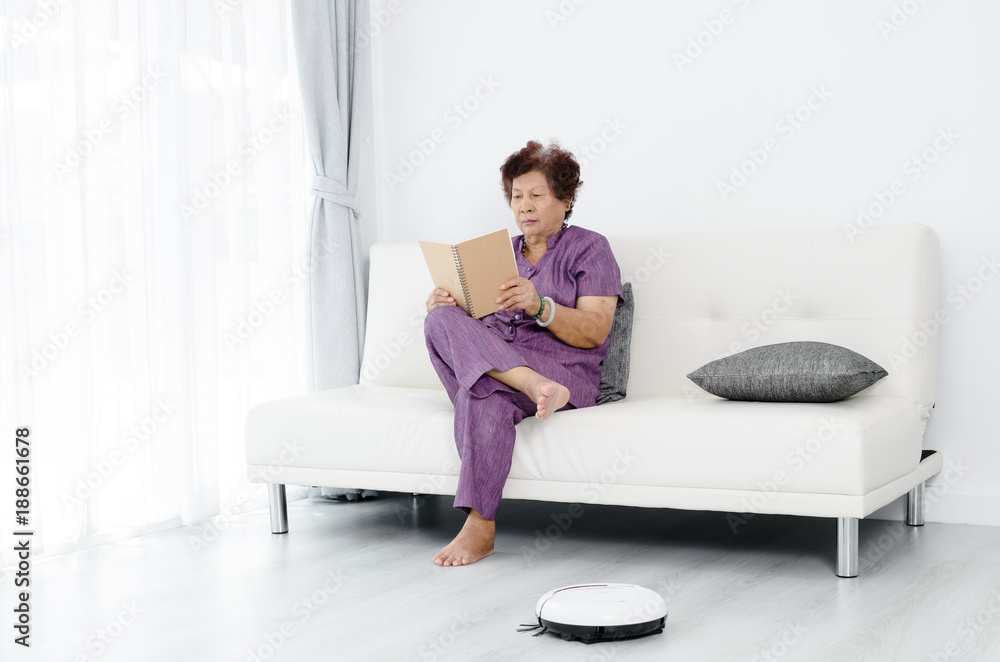 Happy Asian senior woman reading a book at home, cleaning robot working.