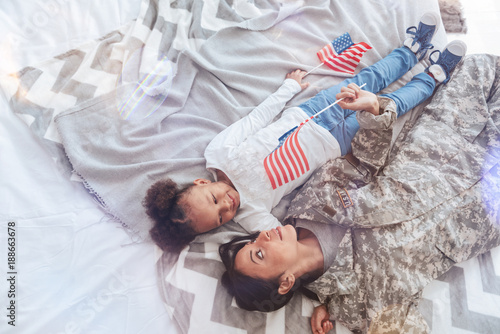 Pleasant relaxation. Nice positive attractive woman holding an American flag and looking at her daughter while lying with her on the bed