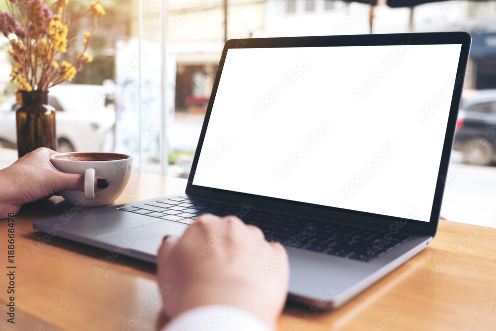 Mockup image of a businesswoman's hand using laptop with blank white desktop screen while drinking hot coffee on wooden table in cafe Mockup image of a businesswoman's hand using laptop with blank white desktop screen while drinking hot coffee on wooden table in cafe