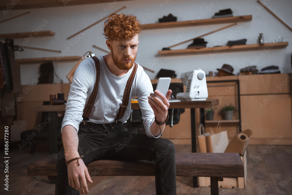 serious young fashion designer holding smartphone and looking at camera in workshop