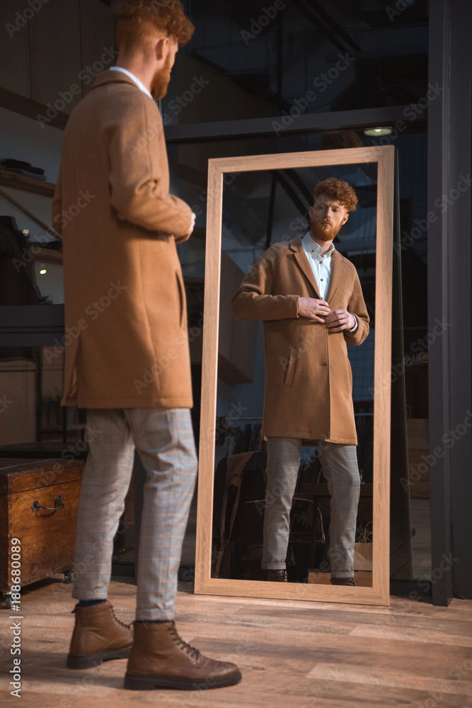stylish young man wearing overcoat and looking at mirror in boutique stylish young man wearing overcoat and looking at mirror in boutique