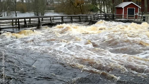 Large amounts of raging water rushes under a wooden walk bridge with icicles on its underside. Red shed beside bridge. The river Morrumsan in southern Sweden.