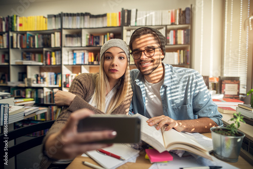 Close up of attractive beautiful hardworking stylish hipster young couple taking a selfie in the library or classroom near the sunny window. © dusanpetkovic1