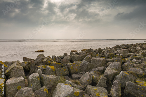 The clouds convey a threatening mood over the calm North Sea at low tide in the Watt. Artificially piled rocks protect the coast from storm surges - Mehldof Bay, Mehldorf, Germany