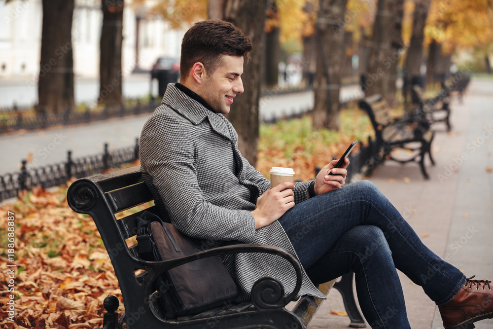 Image of smiling brunette male in coat and jeans drinking takeaway coffee and using his mobile phone, while sitting on bench in park Image of smiling brunette male in coat and jeans drinking takeaway coffee and using his mobile phone, while sitting on bench in park