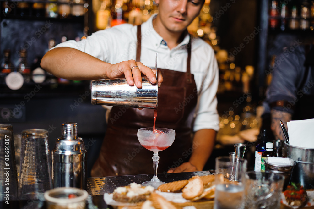 Barman in a brown leather apron pouring fruit alcoholic cocktail into the glass