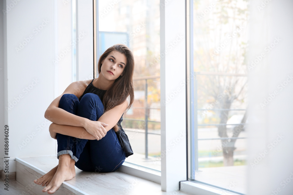 Depressed young woman after breakup sitting on window sill indoors Depressed young woman after breakup sitting on window sill indoors